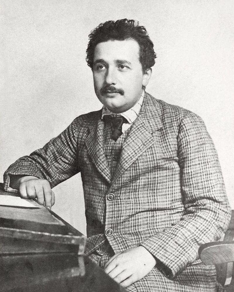 Albert Einstein in his mid-twenties, in a checked suit, seated at a desk and looking off to one side.