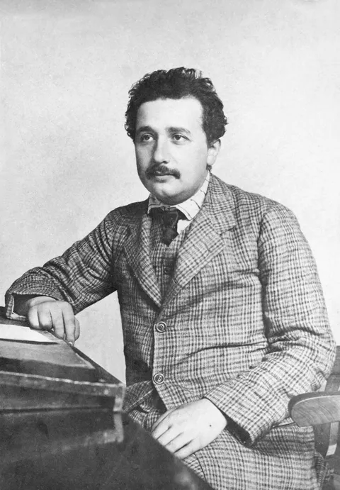 Albert Einstein in his mid-twenties, in a checked suit, seated at a desk and looking to one side.